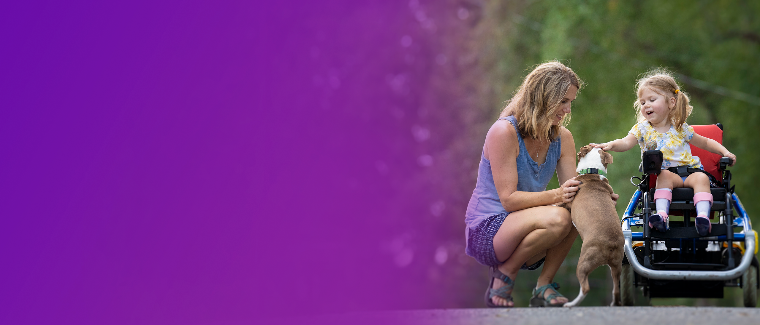 Adalyne, a child with SMA, sitting up in her wheelchair petting a dog. Her mother is crouched down next to her also petting the dog.