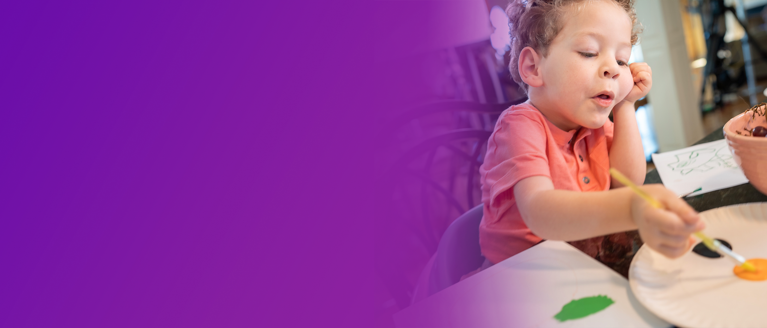 Eli, a 5-year-old with SMA, sitting at a kitchen counter, resting his head in his left hand while mixing paint with a brush in his other hand