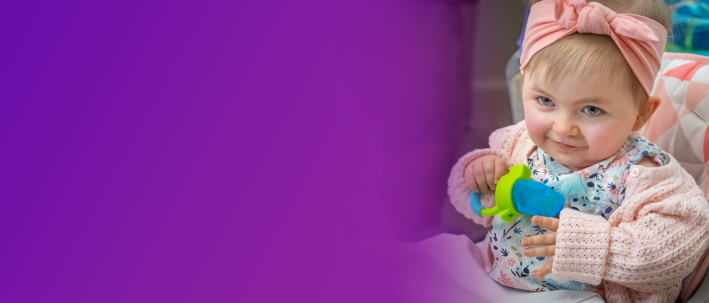 Maisie, a 2-year-old child with SMA, sitting up in a highchair, gripping a green and blue toy