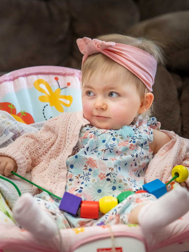 Maisie as an infant, alert and sitting up in her baby carrier. She is playing with blocks and wearing a pink headband